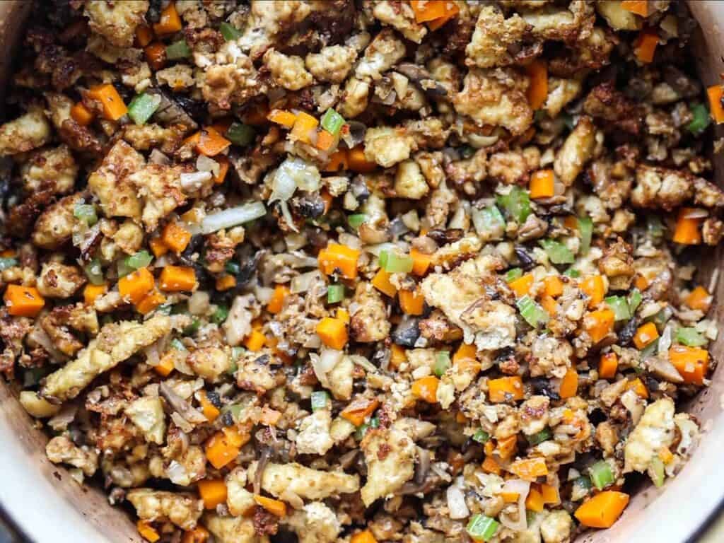 A close-up of a colorful stir-fry with diced carrots, onions, celery, and mushrooms and tofu, mixed together in a large pot.